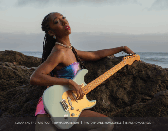 Aviana of Aviana and the Pure Root featured in the 2025 Guitar Girl Magazine calendar pre-planner holding a Fender electric guitar by the ocean