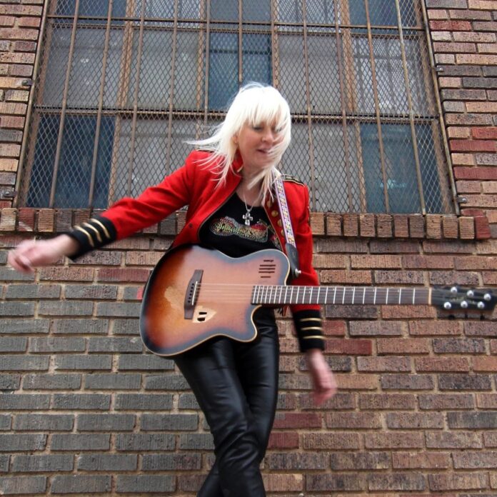 blonde woman posing in front of brick wall with electric guitar