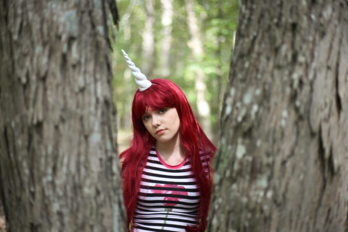 Belle Blue with vivid red hair and a unicorn horn headpiece standing between two trees, symbolizing the imaginative, rebellious energy of her rock persona.