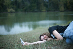 Belle Blue lying on the grass beside a lake, holding a unicorn horn prop, evoking the dreamy, introspective mood of her song “Needed You More.”