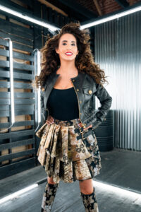 Laura Bryna poses in a black leather jacket and gold floral skirt ahead of her new single “Tornado Weather” premiere on Guitar Girl Magazine