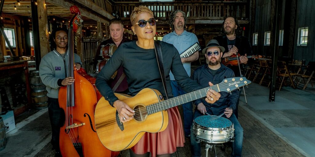Paula Boggs Band poses with acoustic instruments inside a rustic barn venue, featuring Paula Boggs holding a Breedlove acoustic guitar. Photo by Tom Reese.