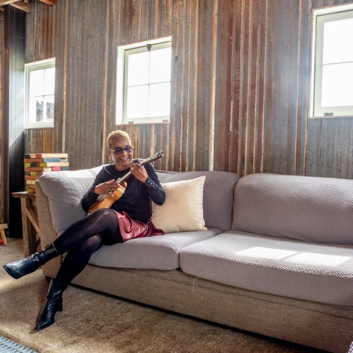 Paula Boggs plays ukulele while seated on a gray couch inside a rustic venue space with natural light streaming through the windows. Photo by Tom Reese.